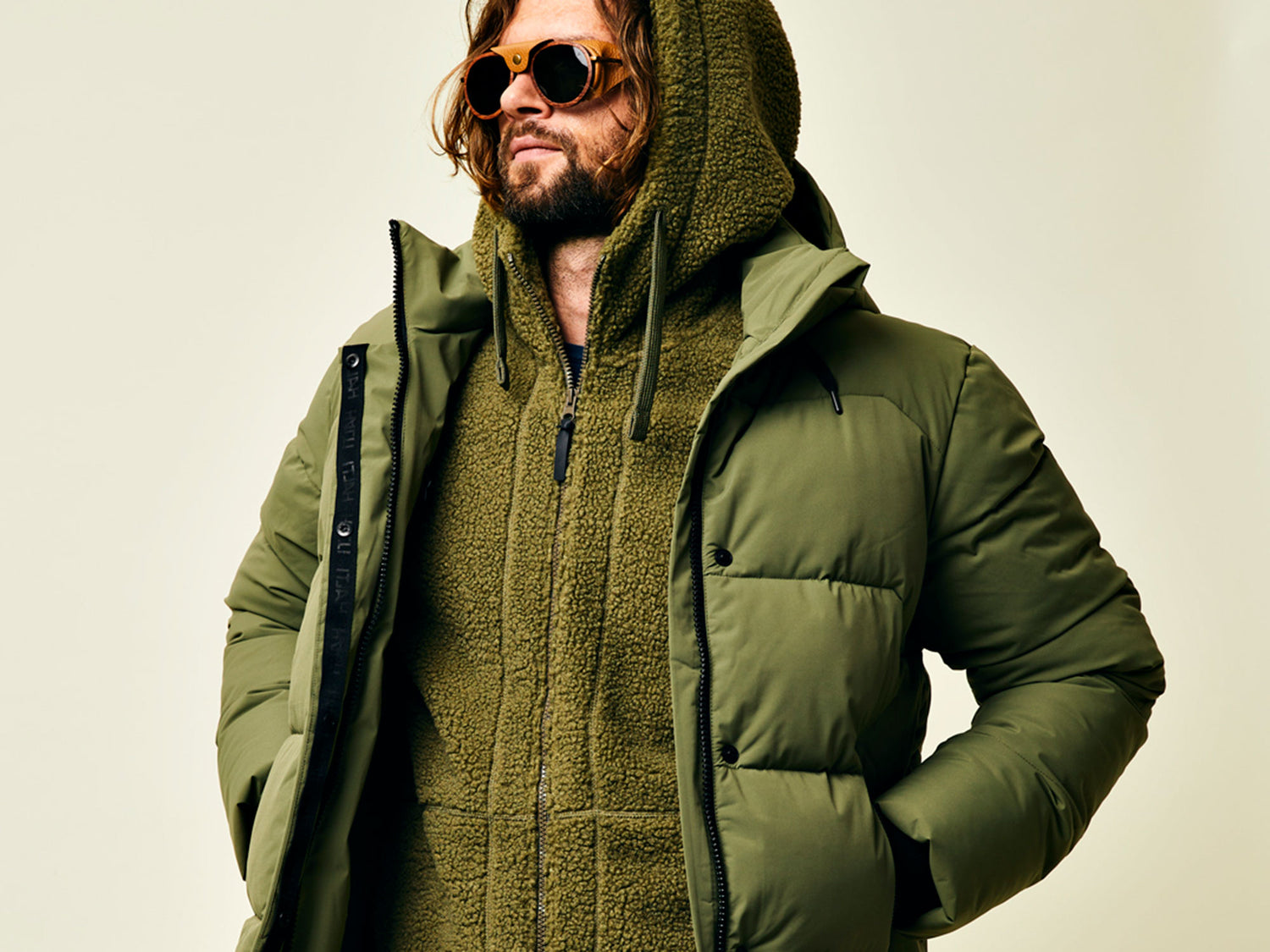 Eine Person, die eine Sonnenbrille, einen grünen Kapuzenpulli und eine grüne Pufferjacke trägt, steht vor einem hellen Hintergrund.