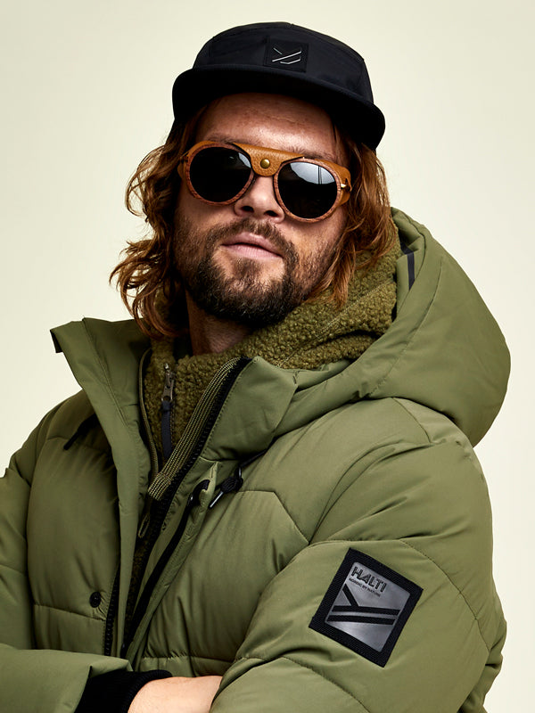 Eine Person, die eine grüne Pufferjacke, eine Sonnenbrille, eine schwarze Mütze und einen grünen Strickschal trägt, steht vor einem schlichten Hintergrund.