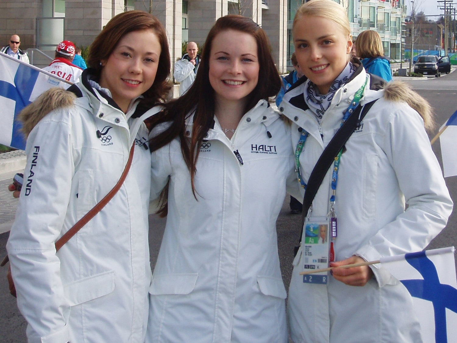 Drei Frauen in weißen finnischen Olympiajacken stehen im Freien, lächeln und halten finnische Fahnen.