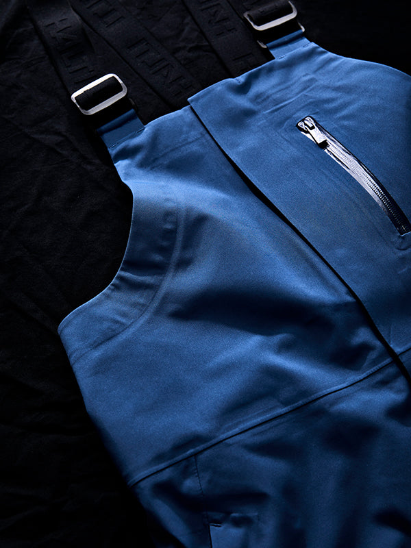 Nahaufnahme eines blauen Overalls mit einer Brusttasche mit Reißverschluss und schwarzen verstellbaren Schulterträgern, flach auf eine schwarze Fläche gelegt.