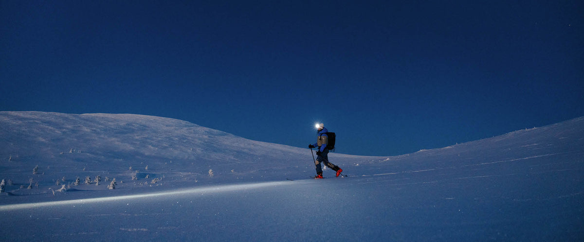 Eine Person mit einer Stirnlampe fährt nachts bei klarem, dunkelblauem Himmel auf einer verschneiten Piste bergauf.