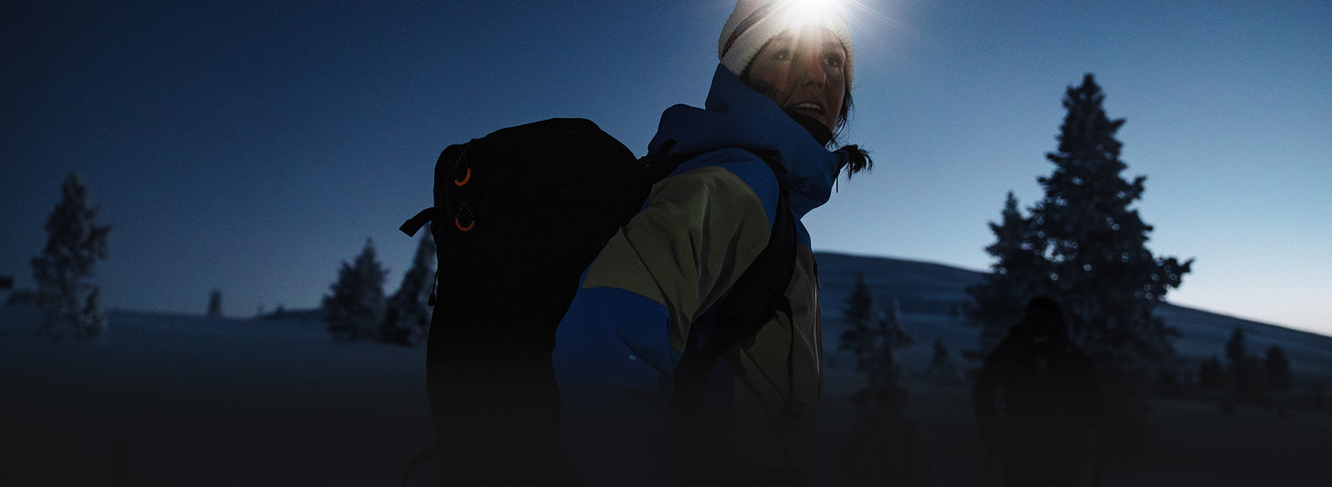 Eine Person mit Rucksack und Stirnlampe wandert in der Abenddämmerung durch eine verschneite Landschaft mit Bäumen und Hügeln im Hintergrund.