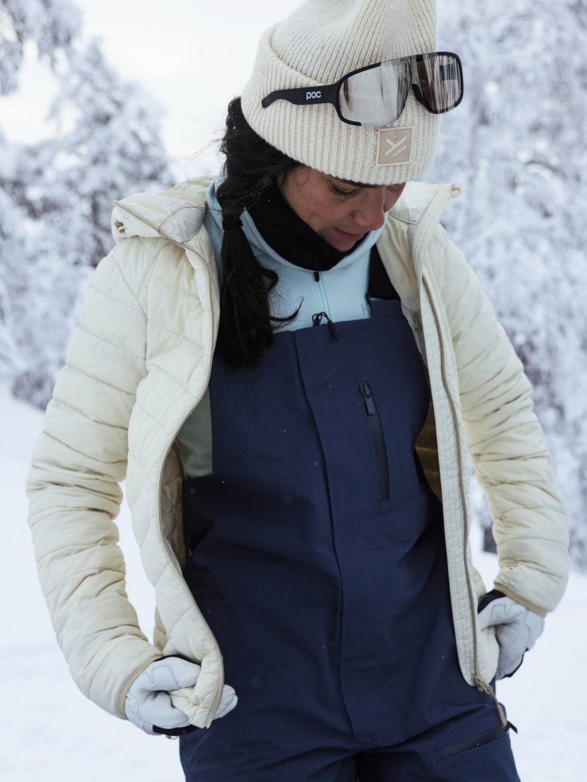Eine Person in einer weißen Halti Huippu Untuvia-Daunenjacke für Damen, einer marineblauen Latzhose, einer Mütze und einer Skibrille steht in einem verschneiten Wald.