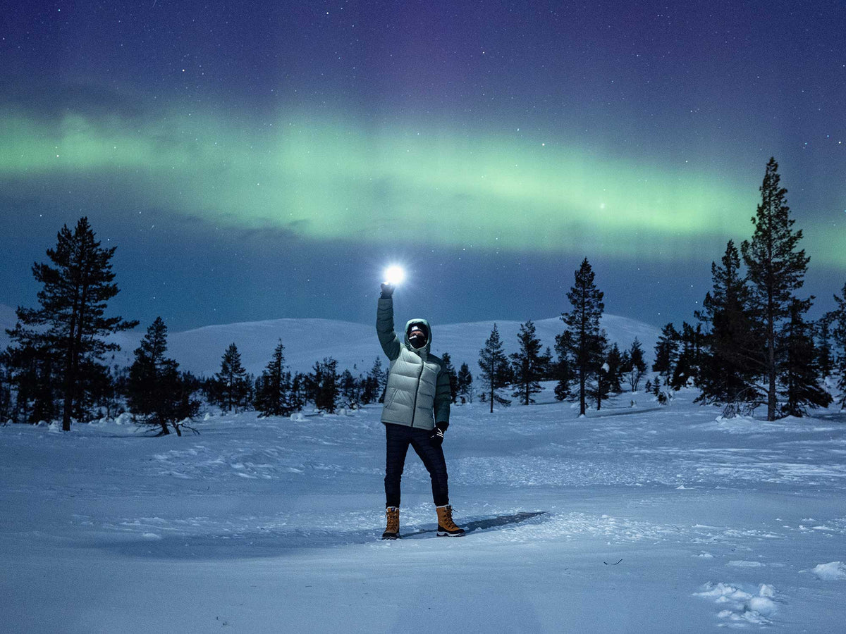 Eine Person steht nachts im Schnee und hält eine Lampe in die Höhe, während das Nordlicht am Himmel hinter ihr zu sehen ist.