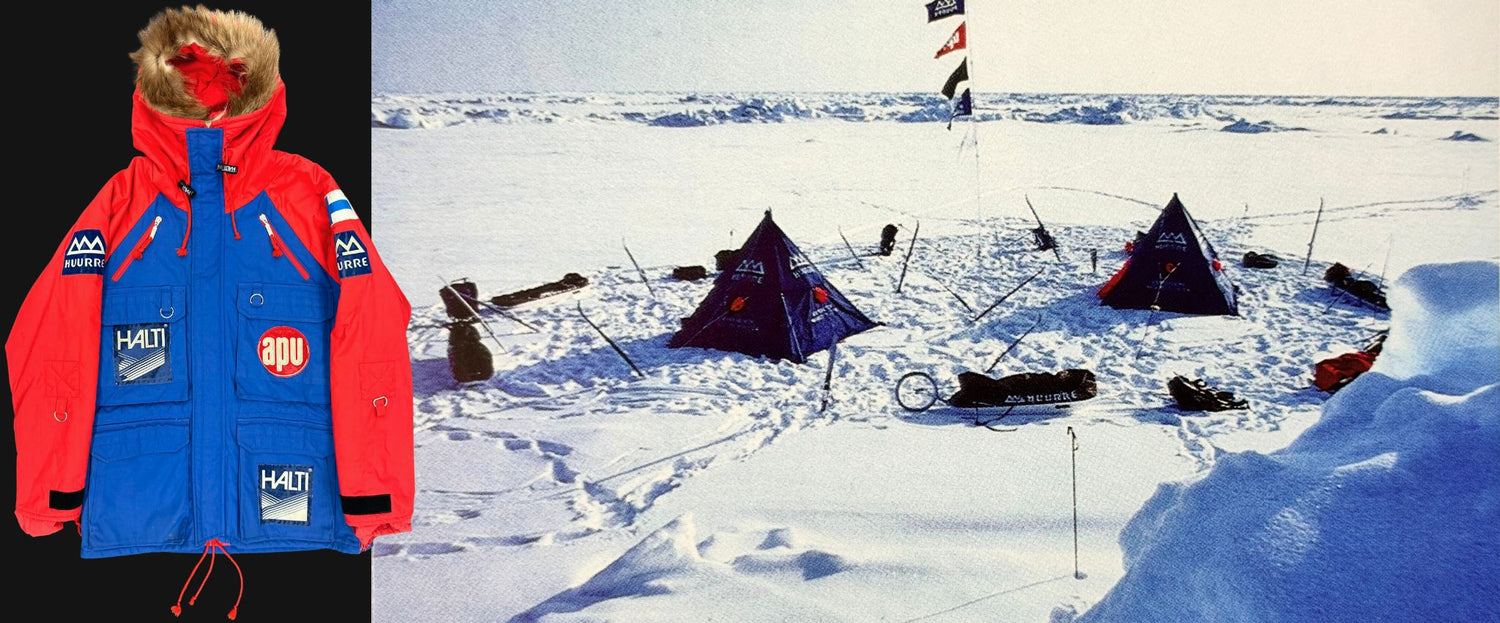 Eine rote und blaue Winterjacke neben einem verschneiten Campingplatz mit blauen Zelten, Schlitten und mehreren Fahnen.