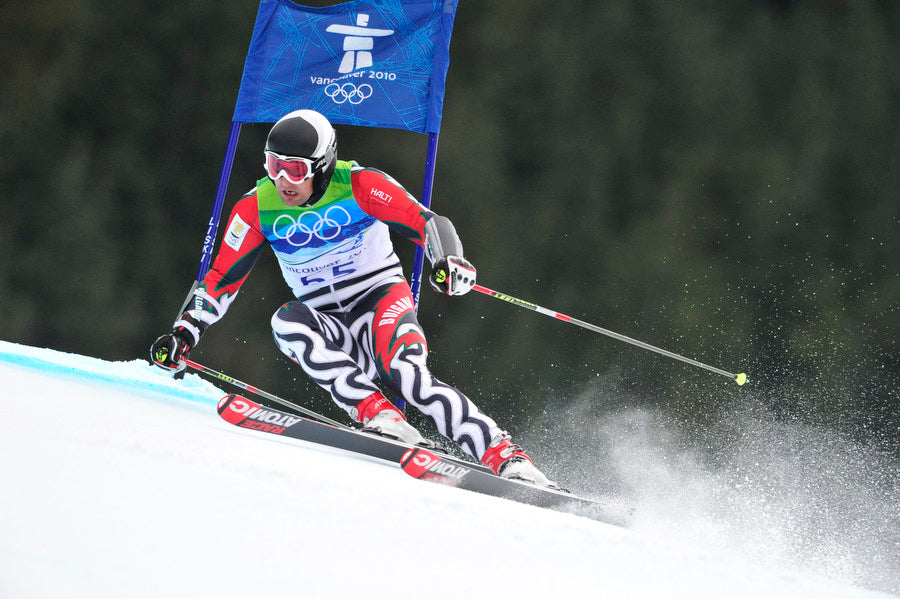 Ein Skifahrer in olympischer Ausrüstung fährt während eines Wettkampfs auf einer verschneiten Piste an einem blauen Tor vorbei.