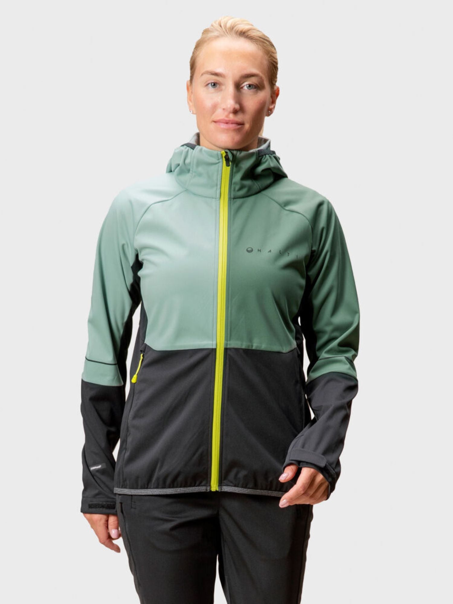 Eine Frau trägt die Halti Exhale Stormwall Jacke in Grün-Schwarz mit gelbem Reißverschluss vor hellem Hintergrund.