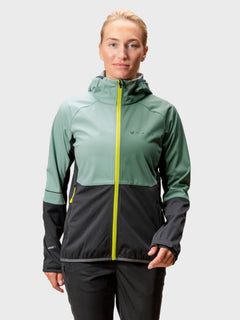 Eine Frau trägt die Halti Exhale Stormwall Jacke in Grün-Schwarz mit gelbem Reißverschluss vor hellem Hintergrund.