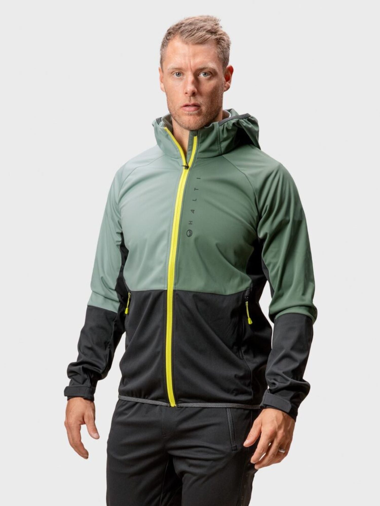 Ein Mann trägt die Halti Exhale Stormwall Jacke Herren mit reflektierenden Details und steht vor einem schlichten Hintergrund.