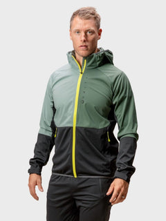 Ein Mann trägt die Halti Exhale Stormwall Jacke Herren mit reflektierenden Details und steht vor einem schlichten Hintergrund.